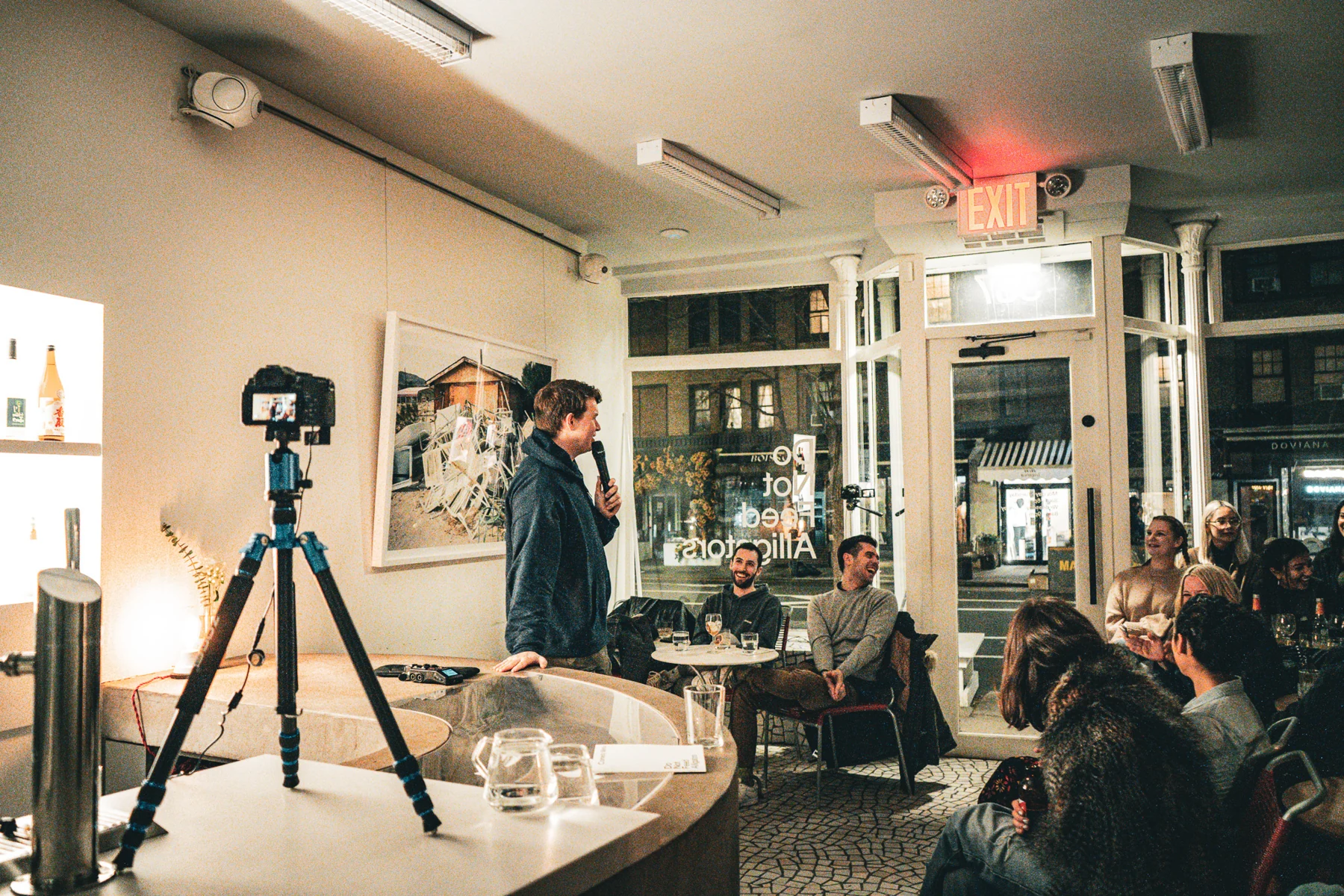 An intimate comedy performance beside a curved counter, with seated audience members and a recording camera.