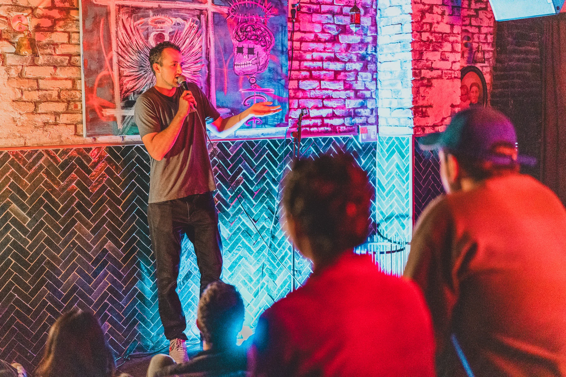 A comedian performing under red and turquoise light in front of a brick wall, with the audience in the foreground.
