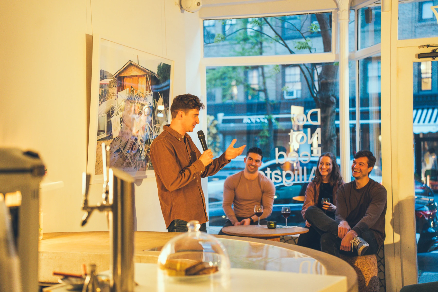 A comedian holding a microphone beside the audience in a warmly lit comedy room.