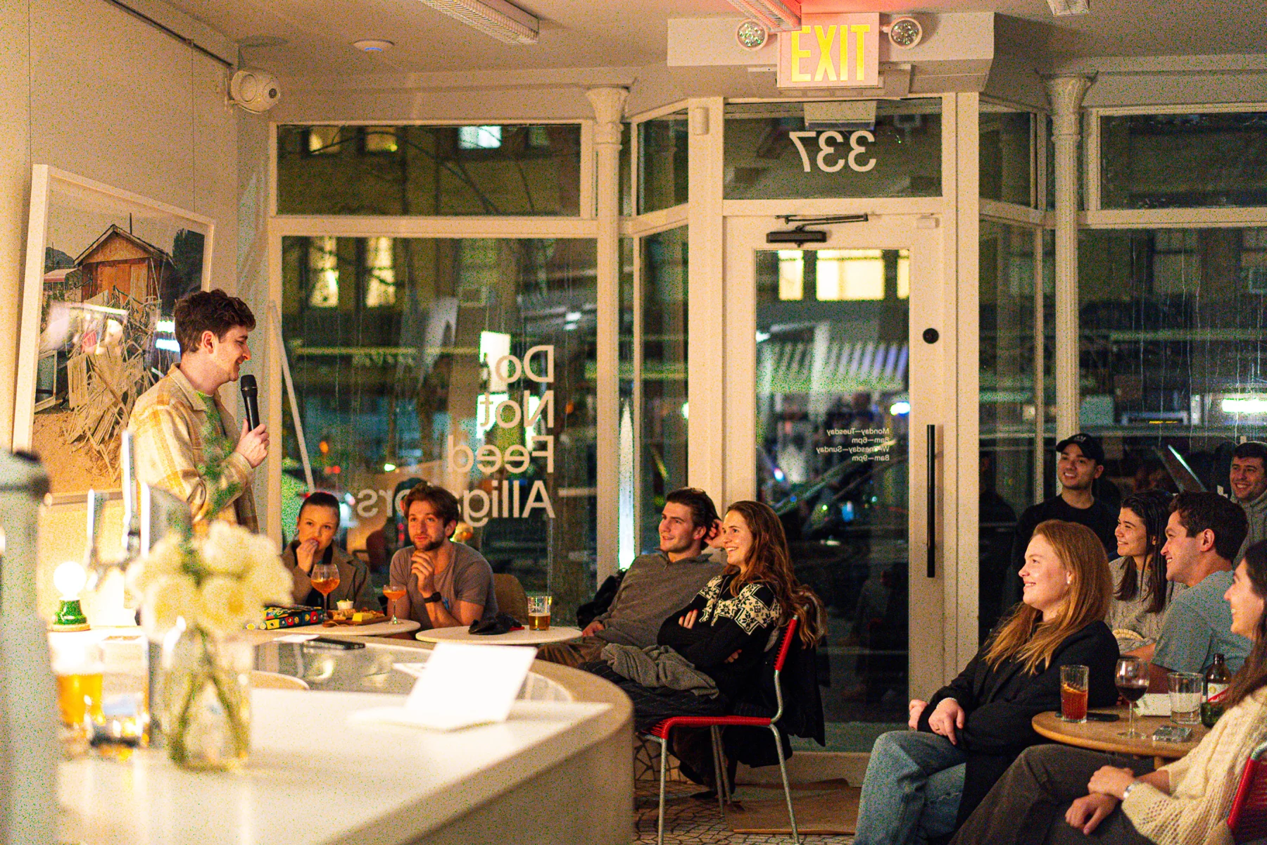 A comedian holding a microphone in a small room with audience members seated around a curved counter.