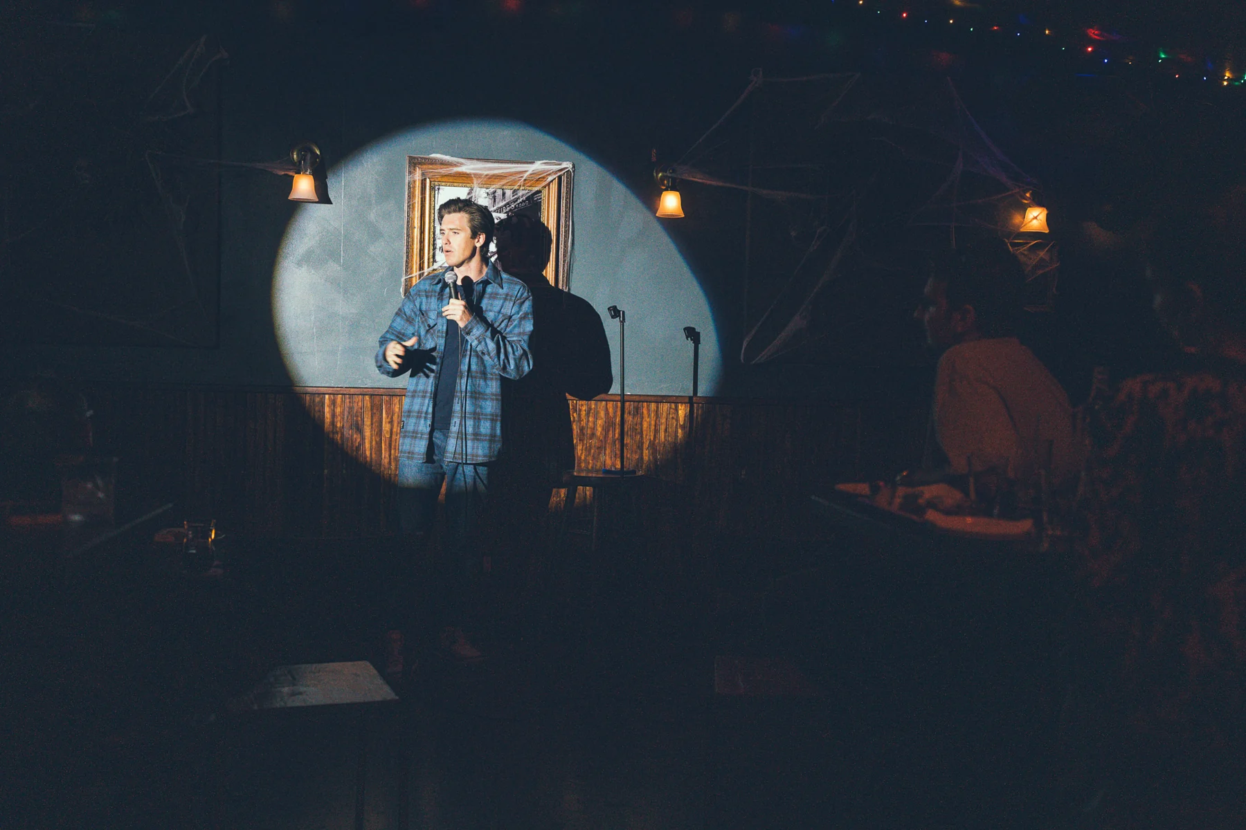 A comedian in a blue checked shirt performing in a circular spotlight, with the audience in the foreground.